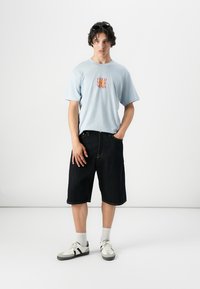 Camiseta de algodón azul claro con texto rojo "ALIMENTA TU MENTE," combinada con shorts de mezclilla oscura. Zapatillas blancas y grises, usando gafas de sol.