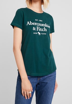 Kobieta w ciemnozielonym t-shircie Abercrombie & Fitch i niebieskich dżinsach, z srebrnymi pierścionkami na lewej ręce.