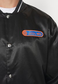 Black satin jacket with snap button closure. Features a colorful embroidered patch with "Puma HOOPS 1973" and basketball graphic.
