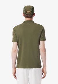 Olijfgroene polo met een kraagontwerp, korte mouwen, en Lacoste-logo op de achterkant; gecombineerd met een bijpassende pet met een ruitpatroon.