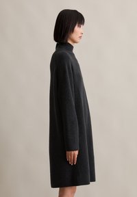 Temno siva pletena obleka, visok ovratnik, ohlapen kroj, dolgi rokavi in ribani manšeti. Gladka tekstura in dolžina do kolen.