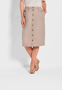 Beige Leinenrock in knieumspielender Länge mit vorderem Knopfverschluss, zwei vorderen Taschen und flachem Bund. Getragen mit hellbraunen Wedge-Sandalen.