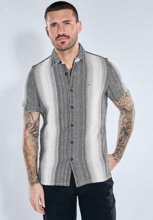Mann mit Tattoos auf beiden Armen, der ein kurzärmeliges, geknöpftes Hemd mit vertikalen Streifen in Grau-, Weiß- und Schwarztönen trägt.