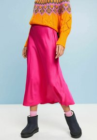 Jupe midi en satin rose avec une silhouette fluide, accompagnée de bottines noires et de chaussettes rose pâle, sur un fond bleu clair.