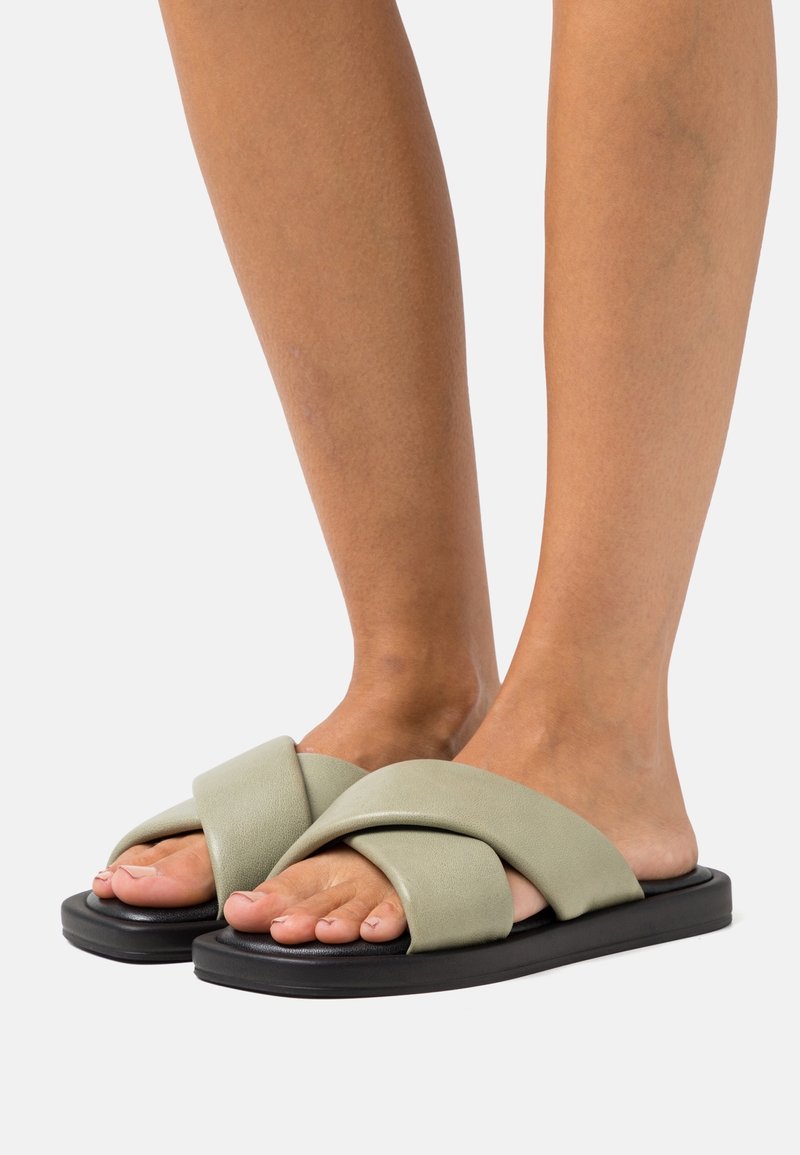 Pieds portant des sandales à enfiler avec des brides larges, rembourrées et croisées de couleur vert clair, ainsi que des semelles plates noires sur un fond neutre.