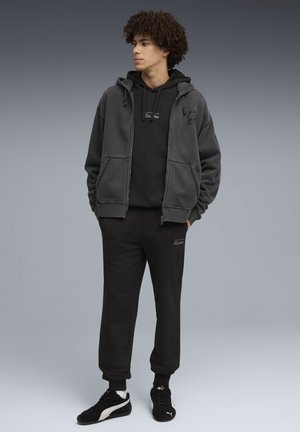 Grauer Kapuzenpullover mit Reißverschluss, getragen über einem schwarzen Kapuzenpullover, kombiniert mit schwarzen Jogginghosen und schwarzen Turnschuhen. Verfügt über schlichte Logodetails.