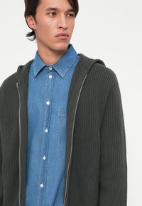 Felpa con cappuccio in maglia grigia scura e texture, con zip frontale e cappuccio, indossata sopra una camicia in denim azzurro chiaro con bottoni e tasca.