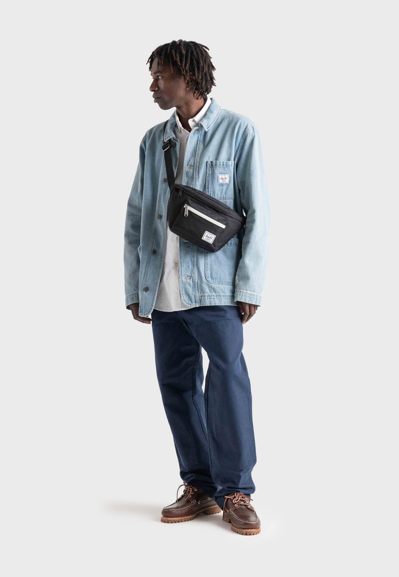 Tenue décontractée comprenant une veste en jean bleu clair, une chemise blanche, un pantalon bleu foncé et des chaussures marron ; inclut un sac bandoulière noir avec des fermetures éclaires.