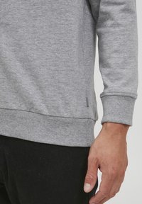 Grå genser med langermet, ribbestrikkede mansjetter og nederste kant. Myk tekstur med en detalj på siden nær den nedre søm.