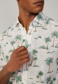 Camisa de lino de manga corta con un fondo blanco y patrones de palmeras verdes, y un cuello abotonado. Tejido texturizado.