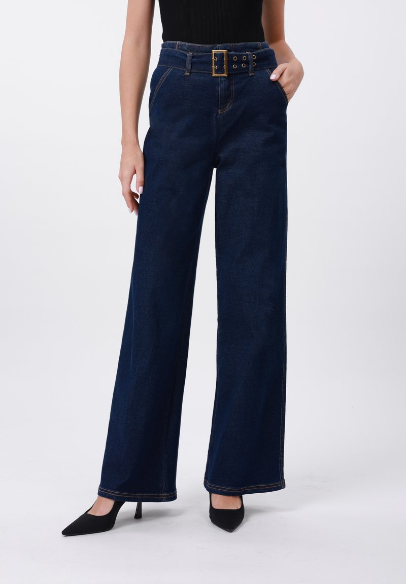Högmidjade mörka jeans med vida ben, med en bältad midja, framfickor och kontrasterande sömmar. I kombination med svarta spetsiga klackskor.