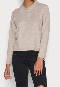 Beige gebreide trui met een kraagnek, drie decoratieve knopen en lange mouwen; gecombineerd met zwarte versleten jeans.