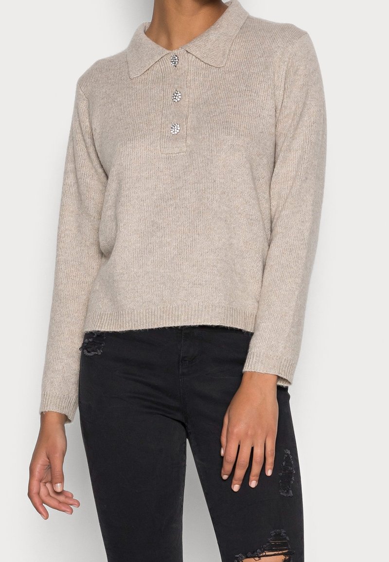 Beige gebreide trui met een kraagnek, drie decoratieve knopen en lange mouwen; gecombineerd met zwarte versleten jeans.