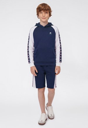 Roodharige jongen die een marineblauwe en witte Champion-hoodie draagt met logostrepen op de mouwen, bijpassende marineblauwe shorts en witte sneakers, staand tegen een witte achtergrond.