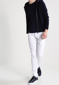 Navy zip-up hoodie over a black shirt, paired with white jeans and navy sneakers. The hoodie features ribbed cuffs and a lined interior.