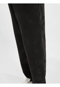 Svarte joggebukser laget av mykt materiale med en tonet logo-design langs siden, med en smal passform og elastisk midje.