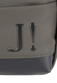 Siva teksturirana torba z gladko črno podlago, ki ima izrazit črno embosiran "J!" logotip. Preprost dizajn, minimalistične akcenti.