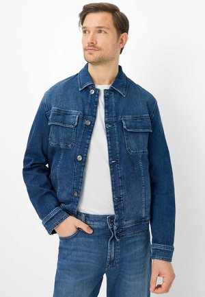 Homme portant une veste en jean bleu foncé et un jean, avec un t-shirt blanc, debout la main gauche dans la poche, regardant légèrement vers la droite.