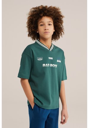 Garçon aux cheveux bouclés portant un tee-shirt de football vert "Bad Boys" et un pantalon bleu, debout avec une main dans la poche contre un fond uni.