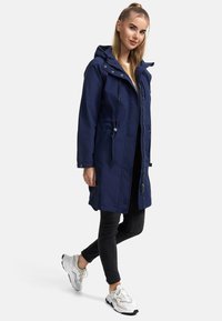 Mörkblå parkajacka med huva, framdragkedja, dragsko och sidofickor. Kombinerad med svarta smala jeans och vita sneakers.