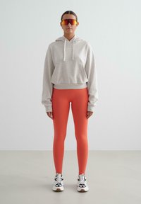 Mujer de pie, vistiendo una sudadera corta beige, leggings naranja brillante, zapatillas blancas y gafas deportivas rojas reflectantes, sobre un fondo liso.