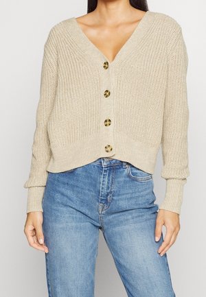 Femme portant un cardigan beige en maille boutonné avec des boutons en écaille de tortue et un jean taille haute bleu, les mains détendues le long du corps.