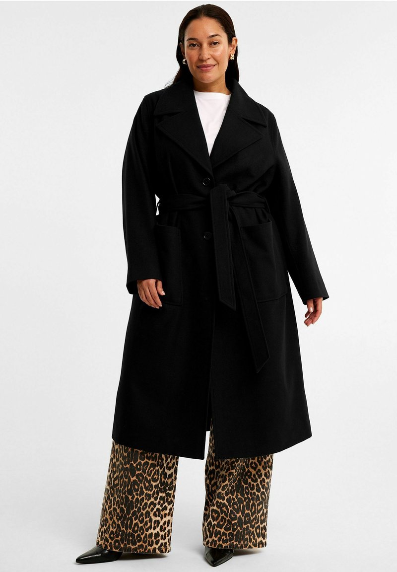 Zwarte lange jas met een riem, grote kraag en twee voorkeuzes. Draag het over hoge taille broek met luipaardprint en zwarte hakken.