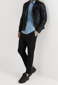 Giacca biker in pelle nera, camicia in denim azzurro chiaro, pantaloni neri a sigaretta e sneakers nere. Presenta tatuaggi sulle braccia e cuciture decorative.