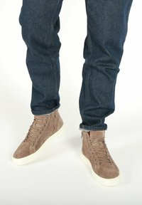 Højtopsneakers i taupe ruskind med flade snørebånd, hvide gummisåler og sidelukninger, parret med mørke denimjeans.
