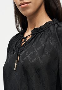 Μαύρη μπλούζα με υφασματικό μοτίβο, με βολάν στο ντεκολτέ και κλείσιμο με κορδόνι. Εντυπωσιακές χρυσές λεπτομέρειες στο στήθος.