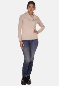 Maglione beige con collo a cowl realizzato in tessuto morbido, dotato di maniche lunghe, abbinato a jeans blu scuro e stivali neri con lacci.