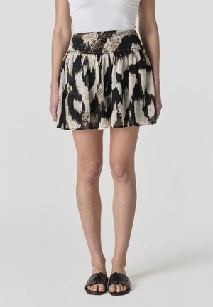 Mujer con falda plisada de patrones en negro y beige, top blanco y sandalias negras con tachuelas, de pie sobre un fondo blanco.