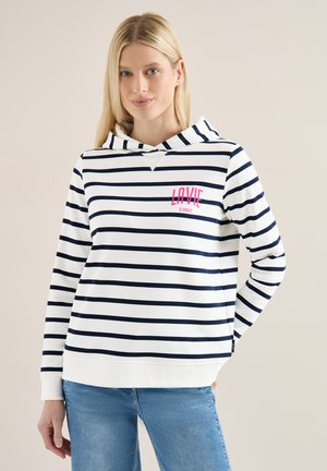 Femme blonde portant un sweat à capuche blanc avec des bandes noires et l'inscription "LA VIE À PARIS" en rose, associée à un jean bleu clair, debout devant un fond beige.