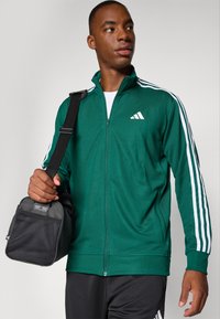 Zaļš rāvējslēdzēja jaka ar baltām svītrām uz piedurknēm, ar Adidas logotipu. Modelis tur melnu somu ar acs priekšējo paneli.