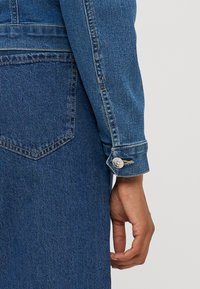 Detalj av en jeansjacka som visar ett mörkblått tyg, knäppt manschett och vikt ärm. Bakfickan har en spetsig design med synlig söm.