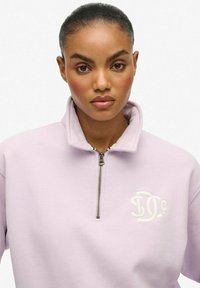 Jasnofioletowa bluza z zamkiem błyskawicznym i wysokim kołnierzem, z białym haftowanym logo na piersi oraz miękkim, teksturowanym materiałem.