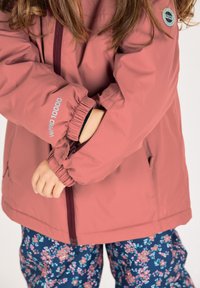 Rosa wasserdichte Jacke mit elastischen Bündchen, Reißverschluss und Seitentaschen, getragen über Blumenmuster-Hosen mit einem marineblauen Hintergrund.