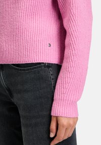 Roze geribbelde gebreide trui met lange mouwen; heeft een textuurpatroon en metallic details nabij de zoom. Gecombineerd met zwarte jeans.