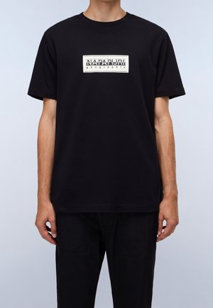 Person wearing a black short-sleeve T-shirt with a white rectangular "Napapijri geographic" logo on the chest, paired with black pants.