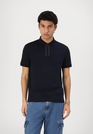 Marineblå polo skjorte med glidelås krage og korte ermer. Har en kontraststripet detalj på kragen. Kombinert med blå denim bukser.