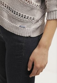 Pull en maille gris clair avec des motifs en tricot ouvert et un ourlet côtelé, associé à un jean foncé, mettant en avant une étiquette de marque bleue en bas.