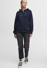 Navy blue hooded sweatshirt with a half-zip front, front kangaroo pocket, and adjustable drawstrings. Paired with cropped black jeans and sneakers.