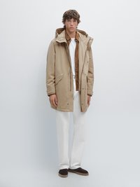 Beige parka met een capuchon en meerdere zakken, gedragen over een bruine shirt en witte broek. Model staat tegen een lichte achtergrond. Bruine schoenen.