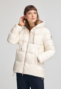Chaqueta acolchada crema con capucha, con herrajes dorados, textura acolchada y acentos de logo. Dos bolsillos frontales con cremalleras.