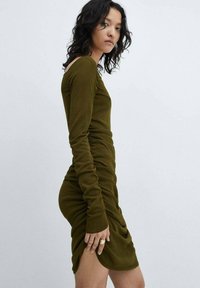 Olivno zelena obleka z dolgimi rokavi z nabrano podrobnostjo ob strani in visokim razporkom, izdelana iz mehke tkanine z gladko teksturo.