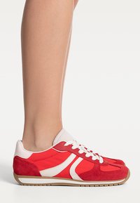Chaussures de sport rouges en daim et tissu, avec des accents blancs dans un design rétro, dotées d'un col rembourré et de lacets. Semelle plate en caoutchouc.