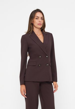 Femme debout portant un blazer marron foncé ajusté à double boutonnage avec un pantalon assorti sur un fond blanc uni.