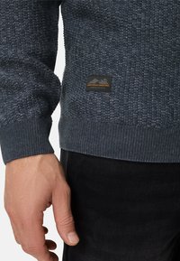 Granatowy sweter o teksturowanej powierzchni z prążkowanymi mankietami i dołem, ozdobiony małą czarną naszywką z pomarańczowym napisem. Materiał wygląda na dzianinowy i trwały.