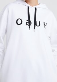 Hoodie bianco con logo stampato in nero. Ha un cappuccio con coulisse nera e punte metalliche. Materiale morbido, vestibilità oversize.