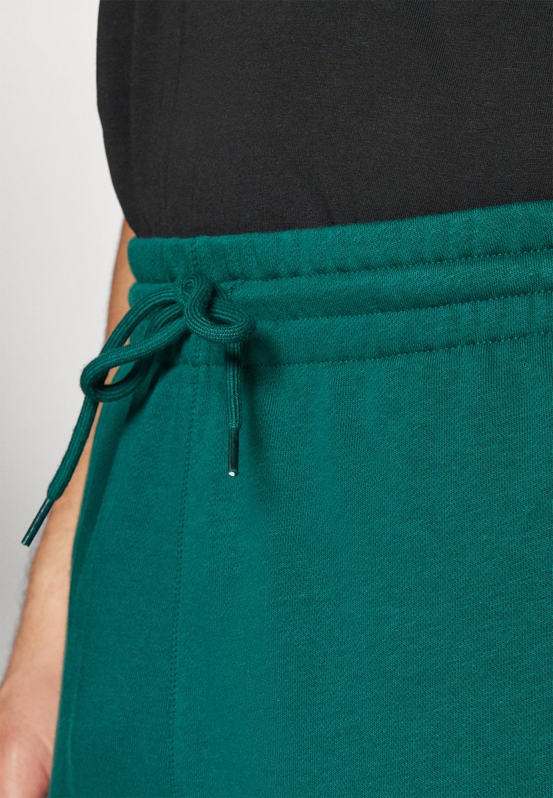 Pantalones de chándal verdes con cintura elástica y cordón. Están hechos de una tela suave y con textura. La camiseta de arriba es negra, contrastando con los pantalones.